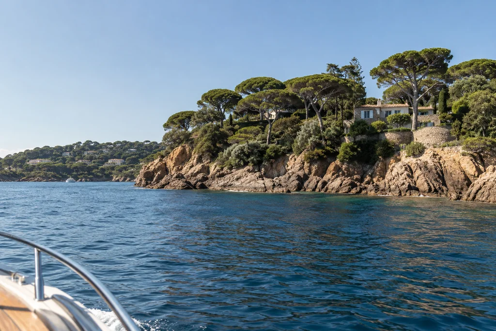 Low pine-covered headlands on the Saint-Tropez peninsula seen from a boat, blue water and warm rocks, no crowded beaches